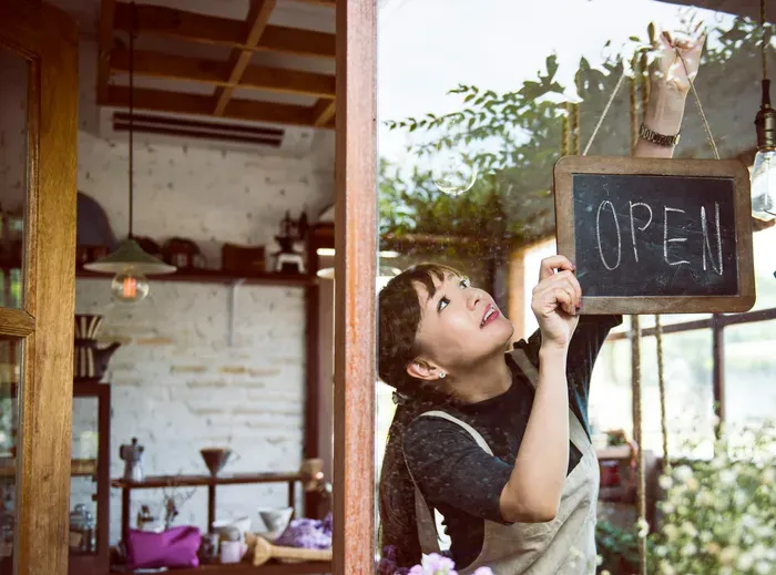Smiling woman hanging an “Open” sign on the door of her cozy café with warm interior and natural light